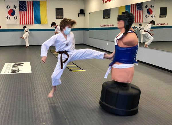 Photo of a boy kicking a Bob martial arts bag. Left leg is extended and pushing the bag, while the right foot is off the ground from his kick.
