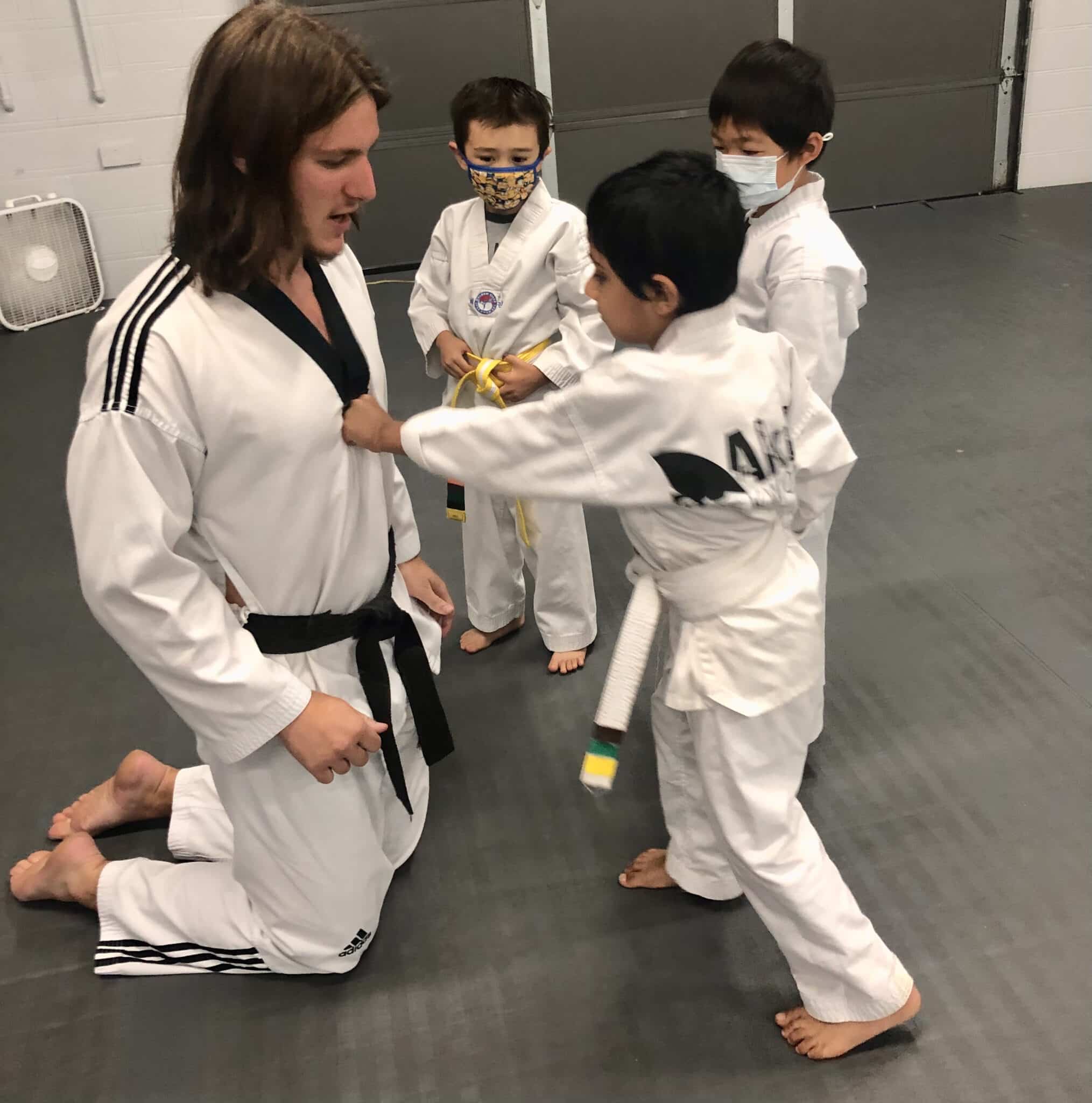 Boy extends his hand in a punch at his Karate teacher's chest while two other boys watch. All are in Taekwondo uniforms from Akula Taekwondo.